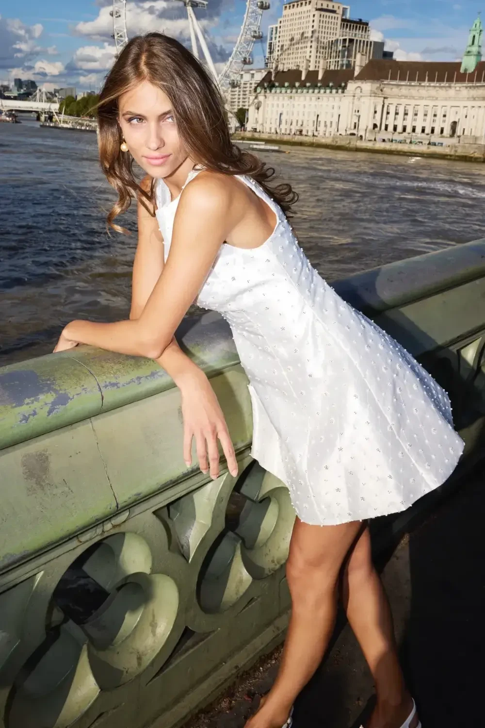 A young woman in a white dress posing by the river with a city landscape in the background.