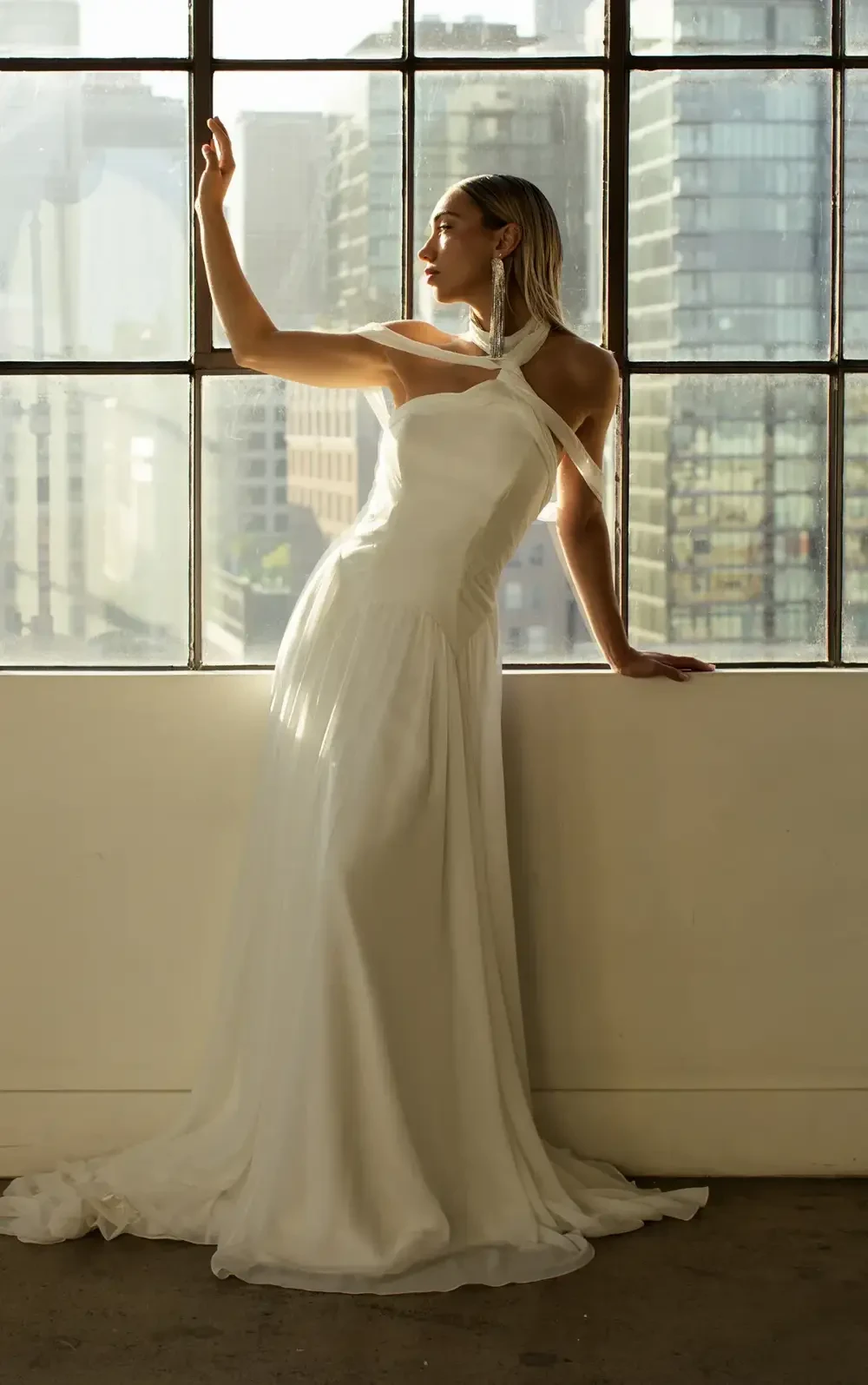 A woman in a white gown stands by a large window, reaching out as sunlight illuminates her features and the room.
