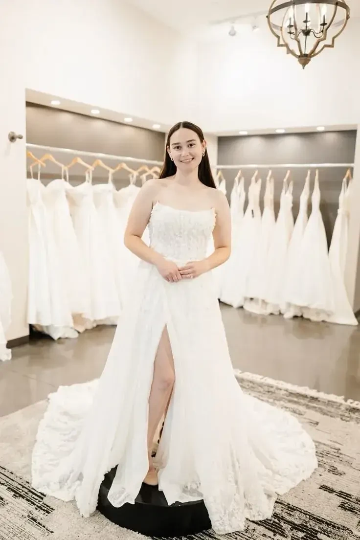 A woman in a white wedding dress is standing on a display platform surrounded by more wedding dresses hanging in the background.
